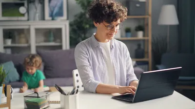 A mother working remotely from home on her laptop while her child plays in the background.