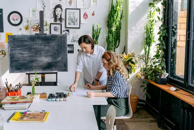 Instructor guiding a student in a vibrant art studio filled with plants and artworks.